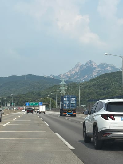 The image depicts a highway scene in Goyang-si, South Korea, with a white SUV in the foreground driving on the right lane. The SUV has a "TUCSON" emblem on its rear. In the middle distance, a blue shipping container truck is in the left lane, and a white box truck is further ahead in the right lane. The highway has multiple lanes and is bordered by a median. In the background, a mountainous landscape with lush green trees and rocky peaks is visible under a partly cloudy sky. Several power lines and transmission towers are interspersed throughout the scene, indicating a developed, semi-urban or suburban area. The overall impression is of a busy highway leading towards a natural landscape.