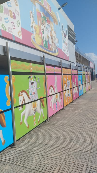 The image captures a brightly colored mural depicting a carousel with various characters on horses, displayed along the exterior wall of a building. The mural is divided into panels, each featuring a different whimsical character riding a white horse, set against vibrant backgrounds of blue, green, pink, and orange. The carousel itself is prominent in the upper portion of the mural, featuring a duck, horses, and a teddy bear. The building's architecture suggests a commercial or entertainment venue, with a modern facade and an outdoor tiled walkway. The scene is set on a clear day with a blue sky visible above. This mural is located in Pylaia Municipal Unit, Greece.