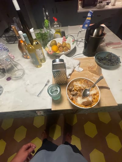 The image depicts a cluttered kitchen counter and the floor below, captured from a first-person, downward-looking perspective, located in a kitchen in Vila Nova de Cerveira, Portugal.
In the immediate foreground, a person's bare feet are visible, standing on a tiled floor composed of brown hexagonal tiles interspersed with yellow hexagonal patterns. The lower part of dark pants or shorts and a light-colored upper garment are also visible, along with the person's right hand on the lower-left side.
The main subject is a white marble or quartz kitchen counter. On a light wooden cutting board, a white round bowl holds a serving of pasta with meatballs in a red sauce, generously topped with grated white cheese. A silver serving spoon rests inside the pasta bowl. Adjacent to the bowl, a silver box grater stands upright, with a folded white and grey checkered kitchen towel draped partially beneath it. A clear glass filled with ice and water is positioned next to the grater, and a silver dinner fork rests on the countertop to the left of the glass. A wedge of hard cheese, consistent with Parmesan, is situated on the cutting board behind the pasta bowl, with scattered white grated particles around it.
Further back on the countertop, several bottles of wine are arranged, including a rosé/sparkling wine bottle and multiple white wine bottles. A clear glass pitcher and a loose purple plastic cap are also present. A decorative white ceramic bowl, featuring painted fruit motifs, contains various fruits such as pears, lemons/oranges, and smaller yellow/brown fruits. Another dark blue/grey bowl holds a quantity of dark, shriveled grapes or berries. A black kitchen appliance, resembling a coffee machine or blender, is situated near the fruit bowl. Other items include a bottle of yellow dish soap with a red pump top and a glass container holding green herbs. A chrome kitchen faucet and a portion of a sink are visible in the background, set against dark grey kitchen cabinets. The scene is illuminated by moderate indoor lighting.