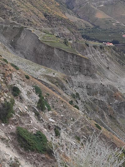 The image depicts a mountainous landscape in Gushal, India. The terrain is steep and rocky, with sparse vegetation consisting of dry grasses and small bushes. In the mid-ground, there is a visible landslide or eroded cliff face with distinct layers of rock and soil. Further in the distance, a settlement is visible with buildings that have red-tiled roofs, and an area that appears to be an orchard or cultivated land. A road or path is visible winding through the mountainside. There are no people or specific actions captured in the scene. The overall environment is rugged and natural, characteristic of a mountainous region.