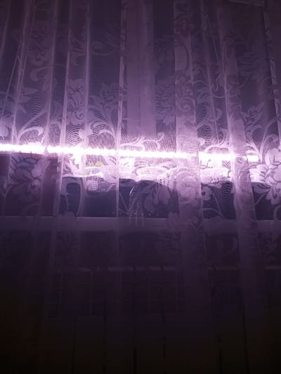 An indoor scene within the city of Яромаска, Russia, is depicted. The primary focus is a window covered by sheer, white or light-colored lace curtains, patterned with floral and scrollwork designs. A prominent, horizontal band of bright pink-purple light originates from behind the curtains, illuminating them from the approximate midpoint of the window. Through the semi-transparent fabric, the window frame and indistinct objects on the windowsill, possibly including a plant or container, are faintly visible. Below the window, in the darker lower portion of the image, a dark, grid-like structure, consistent with a radiator, is discernable through the curtains. No individuals are present. The environment suggests an interior space during low-light conditions, with the artificial light source serving as the main illumination.