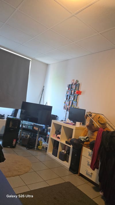 A cluttered room, likely a living space or bedroom, in Vlaardingen, Netherlands. A large black television is centered on a glass-topped entertainment stand, which also holds a black computer tower on the left. To the right of the television, a white shelving unit is filled with various items, including what appears to be a large stuffed teddy bear, several framed pictures or cards, and some storage boxes. A chest of drawers with three visible drawers is positioned to the right of the shelving unit, with dark-colored clothing draped over it. On the floor, a circular beige rug is partially visible, along with a rectangular dark gray rug. The room features tiled flooring and a white drop ceiling. A large window covered by a gray blind is on the left side of the image. The overall impression is one of domesticity with a significant amount of personal belongings.