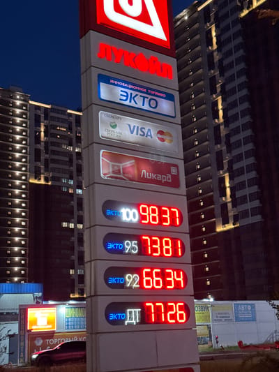 The image depicts a Lukoil gas station sign at night in Leninavan, Russia, displaying fuel prices for various grades. The prices are displayed in red LED numbers: 98.37 for EKTO 100, 73.81 for EKTO 95, 66.34 for EKTO 92, and 77.26 for EKTO Diesel (ДТ). The sign also shows payment options, including VISA, Mastercard, and "Ликард." Nearby buildings are visible in the background. The signs for "SHINOMONTAZH" and "Automotive Spare Parts" can be seen.