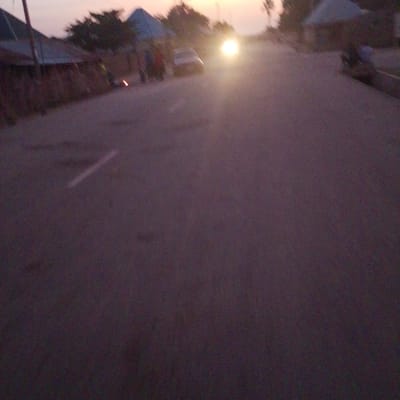 The image depicts a street scene at dusk in Wuro Allahamdu, Nigeria. A vehicle with its headlights on is prominent in the center of the frame, creating a bright glare. Several figures, appearing to be pedestrians, are visible on the left side of the road, some clustered together. To the left, there are structures that resemble buildings or market stalls with conical roofs. The road surface is asphalt, and a white line marking the lane is faintly visible. The overall lighting suggests it is either sunrise or sunset, with the sky showing a pale gradient. The environment appears to be a settlement or village area with some vegetation present in the background.