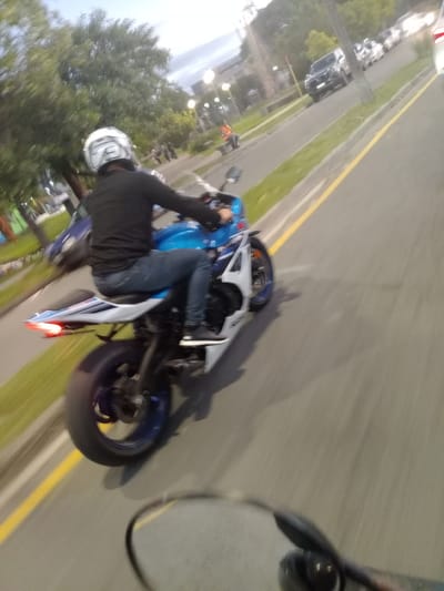 The image captures a dynamic, slightly angled view from a motorcycle in motion, likely in Villa Carlos Paz, Argentina. The primary subject is a person riding a blue and white sportbike. The rider is dressed in a black long-sleeved top and blue jeans, wearing a white and black full-face helmet that obscures their face. Their hands are on the handlebars, suggesting an active riding posture.
The motorcycle itself is predominantly blue on the tank and front fairing, with white sections on the side and rear. A red taillight is visible at the back. The background is blurred, indicating speed and movement. The setting appears to be a paved road or track, featuring a yellow double-line marking on the right side. To the left, there is a grassy strip separating the main thoroughfare from an area with parked cars. Trees are visible further in the background.
The time of day appears to be dusk or night, as indicated by the ambient low light and artificial lights (possibly streetlights or track lighting) glowing in the distance. The overall impression is one of speed and a focused activity of motorcycle riding. No discernible text is visible on any objects or in the background.