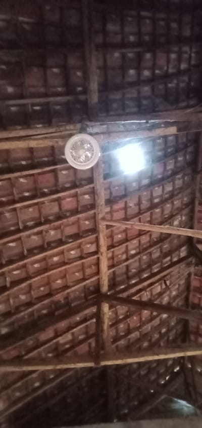 The image depicts a view of a roof structure, likely inside a building. The roof is constructed with wooden beams and red clay tiles. A white ceiling fan is mounted on one of the beams. A bright light source, possibly sunlight, is visible through a gap in the roof. The image was taken from Bulukandang, Indonesia.