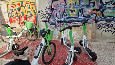 In Faro, Portugal, a row of docked Lime electric scooters and one electric bicycle are parked against a concrete wall adorned with vibrant graffiti. The graffiti features bold, abstract designs in yellow, red, blue, and purple. The ground is a mosaic of red and grey cobblestones. The scooter on the far left is an electric bicycle with a green basket and a "Lime" logo. Next to it is another electric scooter, followed by two more electric scooters. A fifth electric scooter is partially visible on the right. The lighting suggests it is daytime, with shadows indicating a sunny environment. A watermark at the bottom left corner of the image reads "Galaxy S22."