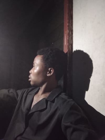 A medium shot captures a young Black person in profile, facing left, sitting indoors against a light-colored wall with a dark doorway to their right. Their skin is illuminated by a strong light source, highlighting their facial features and creating a glossy sheen. They are wearing a black, collared shirt that is unbuttoned, revealing a hint of chest hair. The background is divided between a dark, shadowy area and the brightly lit wall. To the right of the person's head, a distinct shadow of their head and upper body is cast on the wall, mirroring their profile and texture of their hair. The lighting suggests it might be nighttime, or the room is dimly lit with a directional light. There is no visible text in the image. The setting appears to be a simple interior space, possibly a room in Orodo, Nigeria. The person's gaze is directed away from the camera, and their expression is neutral, offering no specific emotional cues. The overall mood is contemplative, with the strong chiaroscuro effect adding a dramatic quality to the scene.
