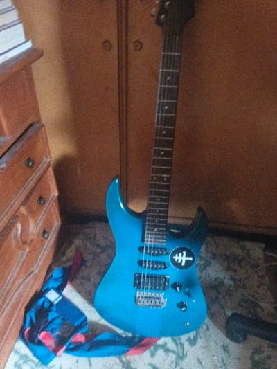 The scene depicts an indoor setting within the city of Dogsenang Gorphoog, Bhutan. A metallic blue electric guitar, featuring a black headstock, dark fretboard, multiple pickups, and a tremolo bridge, leans against a brown wooden wardrobe. A circular black decal with a white stylized key or cross symbol is affixed to the guitar's body. To the left, a multi-drawer brown wooden dresser with dark circular knobs is partially visible, topped by stacked light-colored objects, possibly books. A crumpled fabric item displaying blue, red, and black patterns lies on the floor near the base of the dresser and guitar. The floor surface exhibits a light-colored, mottled pattern. A small black object is partially visible on the floor to the right of the guitar's base. No individuals are present.