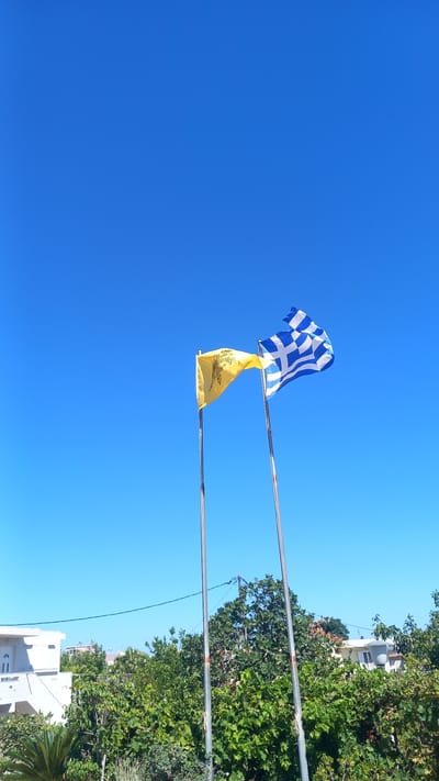 The image shows two flags flying on flagpoles against a clear blue sky. The flag on the right is the national flag of Greece, with its characteristic blue and white stripes and a cross. The flag on the left is yellow and features a black double-headed eagle symbol, likely representing an ecclesiastical or historical Greek flag. Both flags are illuminated by sunlight. Below the flags, lush green trees and foliage are visible, with hints of buildings and structures peeking through the vegetation, suggesting an outdoor setting in a populated area. The overall impression is a sunny day, likely in Greece, given the presence of the Greek flag and the general environment. The location is specified as Platani, Greece.