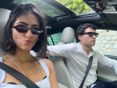 Two people are in the front seats of a car with an open sunroof. The driver is a male with dark curly hair, wearing sunglasses and a vertically striped button-down shirt, with a black seatbelt. The passenger is a female with dark hair, wearing sunglasses and a white top with a necklace, also wearing a black seatbelt. The scene appears to be located on a road near Èze, France, due to the visibility of trees and foliage through the sunroof and side windows.