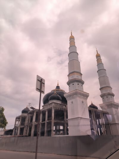 The image displays a large mosque building, which appears to be under construction or recently built, viewed from a low angle, likely from a vehicle. Two prominent and ornate minarets dominate the right side of the frame, reaching high towards a heavily overcast sky. These minarets are primarily white, featuring intricate textured patterns and gold-colored bands and finials at their peaks.
The main mosque structure, partially obscured by the minarets and a foreground sign, shows exposed concrete pillars and beams, indicating ongoing construction. Several dark-colored domes, also topped with golden finials, are visible on the building's roofline. Scaffolding can be seen on the left side of the main structure.
In the foreground, a blurry white and brown road sign on a pole partially obstructs the view of the mosque. Below the construction site, a grey corrugated metal fence runs horizontally, separating the property from a dark road. Faint reflections, consistent with being viewed through a car window, are visible in the lower right corner. A small patch of green foliage is visible on the far left.
The sky is filled with thick, greyish-pink clouds, suggesting an overcast daytime. The scene captures the construction of a grand religious edifice in Keudematangglumpangdua, Indonesia. No clear text is discernible in the image.