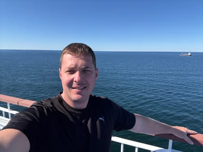 A selfie is taken by a man with short brown hair and fair skin, wearing a black Puma t-shirt. He is standing on a deck with a railing made of brown wood and white metal. The railing is in the foreground, and the man's left arm rests on it, with his hand in view. Behind him, the vast expanse of the blue sea stretches to the horizon under a clear blue sky. A cargo ship is visible in the distance on the right side of the frame. The scene appears to be a sunny day at sea. The location is likely on the water near Karlskrona, Sweden, as indicated by the presence of a cargo ship, which is common in the shipping lanes around this coastal city.