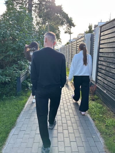 The image shows three people standing on a paved walkway next to a grassy area. The walkway is made of light-colored rectangular pavers. To the left of the walkway is a dense green bush, and beyond that is a tall tree with a slender trunk and dense foliage. To the right of the walkway, there is a wooden fence.
In the foreground, a person wearing a black long-sleeved shirt and black pants is viewed from behind. Their head is turned to the left, with a portion of their short hair visible. To their right, a young woman is also viewed from behind. She is wearing a white long-sleeved sweatshirt and black pants. Her long brown hair is tied back and adorned with a small sparkly accessory. Her body is angled slightly away from the camera. Further in the background, a third person, partially obscured by the woman in white, is also visible from behind. They appear to be wearing black pants, and their long dark hair is also visible.
The scene appears to be outdoors, with natural light suggesting it is daytime. The bright sky in the upper left corner indicates good weather, possibly late afternoon or early evening due to the low angle of the sun casting light on the scene. The presence of the fence and walkway suggests a residential or park setting. The overall mood is casual and relaxed, with the individuals seemingly walking or standing together. There is no visible text in the image.