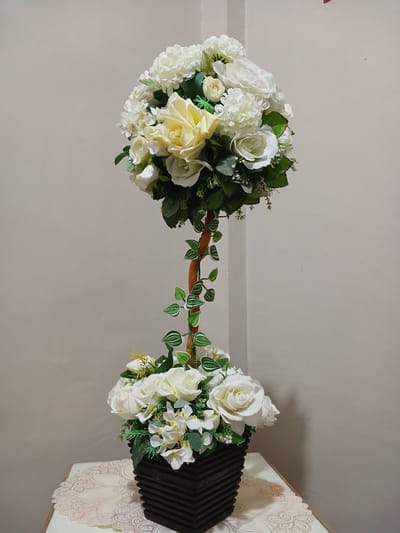A close-up, eye-level shot captures an artificial floral arrangement in a dark brown, square vase. The arrangement features a prominent sphere of white and cream roses and hydrangeas at the top, supported by a slender wooden stem with green vine accents. Below the sphere, a smaller bouquet of white roses and delicate white flowers rests in the vase, which sits on a patterned tablecloth. The background is a plain, light-colored wall. The lighting is neutral, creating soft shadows. The location is Geudonggeudong, Indonesia.