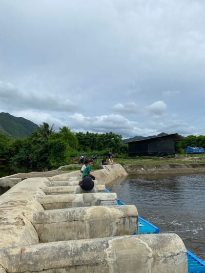 The image depicts a concrete structure, possibly a small dam or spillway, with water flowing through it. Several children are seated on the edge of the structure, appearing to be fishing. In the background, there is lush green vegetation, including trees and a distant mountain range. Several motorcycles are parked near the bank, and a simple, dark-colored structure with a slanted roof is visible further in the distance. The sky is overcast with clouds. The overall scene suggests a rural or semi-rural environment in Aneuk Paya, Indonesia, where children are engaged in leisure activities by the water.