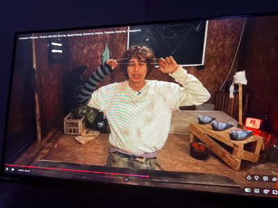 The image shows a screen displaying a video. The video appears to be a scene from a Russian-language YouTube video, as indicated by the text overlay in Cyrillic. The video's title is visible at the top of the screen: "6 Комнат! Обмани - Получи 500.000! (Бустер, ФреймТамер, Кокошка, Горилла, Куертов, Парадеич)". This translates to "6 Rooms! Deceive - Get 500,000! (Buster, FrameTamer, Kokoshka, Gorilla, Kuertov, Paradyich)". The scene depicts a person with curly hair, wearing a white t-shirt with horizontal stripes and a grey and black striped sleeve on their left arm, stretching a rubber band across their eyes with their hands. They are standing in a dimly lit room with wooden walls and floor. In the background, there is a bed, a wooden crate, and a chalkboard with drawings, including an eye within a triangle and a bird within a triangle. There are also several pigeons on a wooden structure in the foreground. The timestamp at the bottom left indicates the video is at 1:30:26 of 2:37:27. The video player controls are also visible. There is no visual information in the image that would indicate the location within the city of Kuvshinovo, Russia.