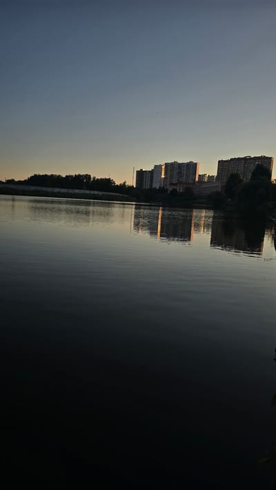This is a rotated photograph of a waterfront cityscape at dusk. The scene is dominated by a body of water on the right, reflecting the sky and buildings on the left. Tall, modern residential buildings, characteristic of a city, line the left bank of the water. The setting sun casts a warm, golden light on the facades of these buildings, and their reflections are visible on the rippling surface of the water. The sky is a gradient of pale blue and yellow, indicating the late hour of the day. Trees line the edge of the water, partially obscuring the lower levels of the buildings and creating a natural buffer between the urban structures and the water. The overall mood is serene and peaceful, with the stillness of the water and the soft lighting contributing to a tranquil atmosphere. There is no visible text in the image. The location context is Ashcherino, Russia.