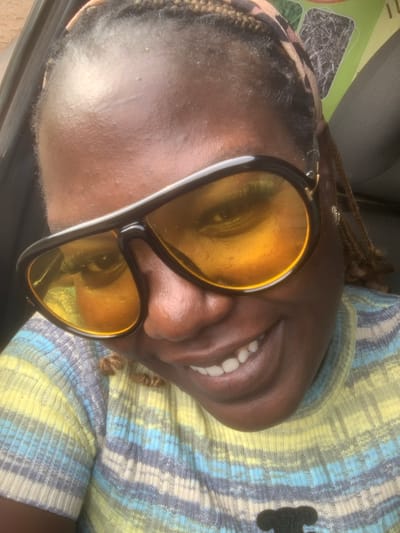 A close-up, high-angle, outdoor selfie captures a Black woman with braids, wearing large, dark-rimmed sunglasses with yellow lenses, and a striped, multicolored, ribbed knit sweater. She is smiling, revealing white teeth, and her head is tilted slightly to her left. The background shows a hint of a green and white sign, possibly indicating a product or location, and the interior of a vehicle, suggested by the presence of a car door and seat. The lighting suggests it is daytime. The location is described as Assob Hausa, Nigeria.