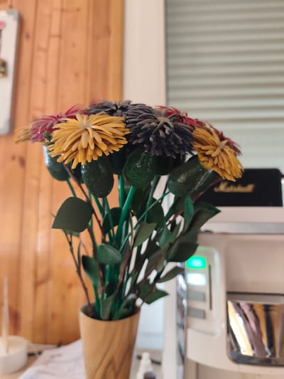 A bouquet of artificial flowers, featuring yellow, red, and dark grey blossoms, is displayed in a light brown wooden vase. The flowers have green stems and leaves, some of which appear to be avocado-shaped. In the background, a light wood-paneled wall is visible, along with a white appliance with a green illuminated indicator. A black object with the gold lettering "Marshall" is also present on the appliance. The scene appears to be indoors, possibly in a domestic or commercial setting. No people are visible in the image. The location is not specified as Edineț, Moldova.
