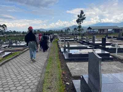 The image shows a paved walkway in a cemetery located in Nzulo, Democratic Republic of Congo, at coordinates -1.6 latitude and 29.124 longitude. The walkway is lined with rows of black granite headstones. A group of people, mostly women, are walking along the path, some carrying white flowers. In the foreground, a man in a red helmet is walking toward the camera. Behind him, a small group of people are walking in the same direction. The cemetery is situated on a hillside, with trees and a metal building in the distance. The sky is cloudy with a few patches of blue.