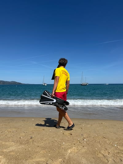 A lone male lifeguard, seen from behind, walks on a sandy beach in Le Lavandou, France, towards the turquoise sea under a clear blue sky. He is wearing a bright yellow t-shirt with red lettering that reads "SAUVETEUR LIFEGUARD" and red swim shorts. In his right hand, he carries a pair of black and white swim fins. The gentle waves of the Mediterranean Sea lap the shore in front of him. Two sailboats are visible in the distance on the calm water, along with a distant coastline. The sun casts a long shadow of the lifeguard on the sand.