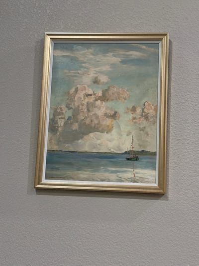 A framed painting hangs on a textured grey wall. The painting depicts a serene landscape with a body of water on the right and a cloudy sky on the left. The water is rendered in shades of blue and grey, with vertical brushstrokes suggesting movement. On the far right edge of the painting, a dark green object with red accents appears to be a small boat or buoy in the water. The sky is filled with large, billowy clouds in tones of white, grey, and pale yellow, with hints of blue peeking through. The overall impression is one of a calm, possibly overcast day. In the bottom right corner of the painting, a signature is visible: "EUGEN BRACHT 1911". The frame is a simple gold-colored molding with a subtle patterned inner edge. The textured wall creates a backdrop that emphasizes the artwork. The lighting suggests it could be daytime, with soft light illuminating the painting and casting a gentle shadow on the wall.