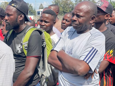 A group of men stand in a crowd in Ville, Democratic Republic of Congo. The men are of African descent and appear to be in their 20s to 50s. The men are wearing a variety of clothing, including a black t-shirt with a red trim, a green backpack, and a white t-shirt with an Adidas logo. The men are looking in different directions. The man in the center of the image is wearing a grey t-shirt with three blue stripes on the right sleeve and an Adidas logo on the left side. He is standing with his arms crossed and looking to the right of the frame. The man is looking to the right. There is another man wearing a white t-shirt with a black and red design with flames and a black shirt underneath it standing to the right of the man in the grey t-shirt. He is looking to the right. The man to the left of the man in the grey t-shirt is wearing a black t-shirt with a green backpack and a red trim on the sleeves. He is looking to the left of the frame. The man to the left of him is wearing a black t-shirt. His head and shoulders are visible. He is looking to the left. The man to the right of the man in the grey t-shirt is wearing a black t-shirt with flames printed on the back. He is wearing a black and red baseball cap. The man to the right of him is wearing a black shirt and a black and red baseball cap. He is looking to the right. The scene takes place in a public space, possibly a street or a market. The men are standing in front of a building with a white wall and a window. There are trees in the background. The sky is clear and blue. The image is likely taken during the day.