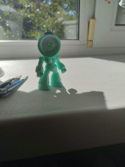 An overhead close-up shot shows a mint green toy robot standing on a white surface near a window. The robot has a round head with a green and orange circular detail on its face. Its body is segmented with arms and legs. To the left of the robot, a pair of blue and silver miniature scissors lies on the surface. The background features a window with a view of green foliage and a bright sky, suggesting an outdoor setting. The scene appears to be indoors, on a windowsill, with sunlight casting a shadow of the robot onto the surface. The location is Rechytsa, Belarus.