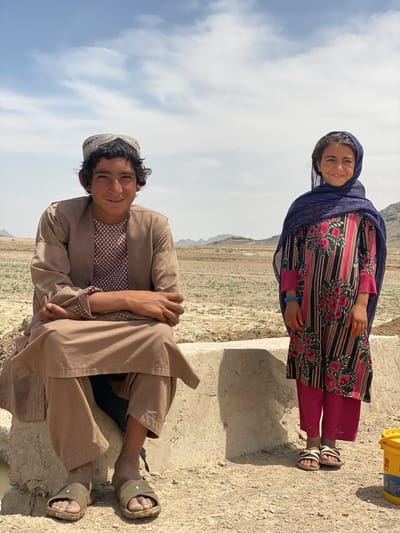 This full-shot photograph captures two young individuals in an arid, rural landscape, identified as Ashoqah, Afghanistan, under a partly cloudy sky. Both subjects are smiling and looking directly at the camera.
On the left, a young man, likely a teenager or young adult, is seated on a low, concrete structure. He wears a light brown, traditional long-sleeved tunic (kameez) over matching loose-fitting trousers (shalwar), and a patterned shirt underneath. On his head, he has a light-colored, intricately embroidered cap. His hands are clasped in front of him, and his feet, appearing dusty, are in simple green slip-on sandals.
Standing to the right of the concrete structure is a young girl, possibly pre-teen or early teenager. She is dressed in a vibrant, long-sleeved tunic-style dress featuring a black and pink striped pattern adorned with large red and pink floral motifs. She wears bright pink trousers underneath and a dark blue headscarf draped over her head and shoulders. Her feet are in light-colored, possibly rope or fabric, sandals. A blue bracelet is visible on her left wrist.
The background reveals a dry, dusty terrain with sparse vegetation and distant mountains under a bright, sunny sky with scattered white clouds. The ground is light brown earth. A small, partially visible yellow bucket with some blue markings is on the ground to the right of the girl. The overall impression is one of candidness and warmth from the subjects in a rugged natural setting.