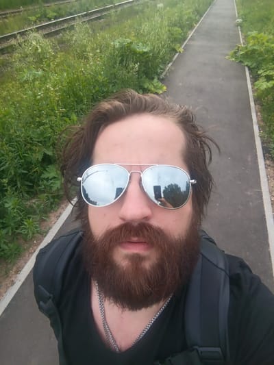 A man with a beard and aviator sunglasses is taking a selfie outdoors. He is wearing a black shirt and a black backpack. The background shows a paved path next to railway tracks and lush green vegetation. The scene is likely in Lesodolgorukovo, Russia, suggesting a rural or suburban setting near a transportation route. The lighting indicates it is daytime, possibly overcast given the reflections in the sunglasses.