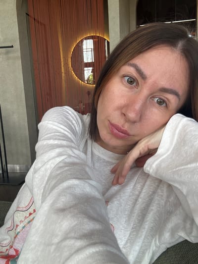 An adult female is captured in a close-up selfie in an indoor setting in Усады, Russia. Her head is slightly tilted, with her left hand resting against her jawline, fingers splayed. She has medium brown hair, hazel eyes looking directly at the camera, and a natural complexion with some subtle skin texture and slight redness on her nose and cheeks. Her lips are softly parted, revealing their natural pink color. She wears a loose-fitting, long-sleeved white top made of a textured, lightweight fabric, showing multiple folds and creases. A subtle silver-colored ring is visible on her left ring finger. A colorful, abstract pattern in red, pink, and white is partially visible on a garment or fabric in the lower left portion of the image.
The background features a decorative wall covered in numerous vertical, reddish-orange to brown strands or ropes. Centered within these strands is a circular mirror, illuminated from behind by a warm, golden glow. The mirror reflects a distinct window or balcony with vertical bars and some green foliage outside. To the far left, a dark, vertical column or frame is visible, along with a dark, horizontal bar fixture. An arched architectural opening is partially visible in the upper right background. The lower foreground shows a textured, green upholstered surface, likely a sofa or chair, on which the subject is seated or resting.