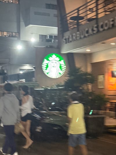 The image shows an outdoor scene at night, likely in a city setting, with a prominent Starbucks logo illuminated by green and white light. In the foreground, several people are visible, some walking and others appearing to sit or rest. Their faces are not clearly discernible due to the low light and motion blur, but their bodies suggest casual attire. One individual is wearing a yellow shirt and blue shorts.

Behind the Starbucks sign, a building with windows and architectural details is visible. The overall lighting suggests it's nighttime, with artificial lights from the establishment and streetlights illuminating the area. The presence of palm-like trees adds to the tropical feel of the location, which is specified as Manila, Philippines. There are no clear indicators of specific weather conditions, but the evening setting is evident.

Partially visible text on a building sign above the Starbucks logo appears to read "STARBUCKS COFF". The atmosphere seems relaxed, with people gathered outside the establishment. The motion blur suggests movement, possibly people walking by or the photographer themselves moving.