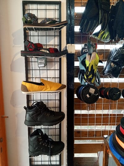 This image depicts an interior retail space in Baza, Myanmar (Burma), featuring various merchandise displayed on wall-mounted racks. On the left, a black metal grid rack with white wire shelves holds five individual footwear items: a pair of brown sandals with patterned green and brown straps on the top shelf, a pair of black sandals with red straps on the second shelf, a single mustard yellow slip-on shoe with a brown heel on the third shelf, a single black hiking boot on the fourth shelf, and another single black hiking boot with laces dangling on the bottom shelf. A product tag hangs below the second shelf of sandals.
To the right, a dark vertical beam separates the left display from another grid display area. A white electrical switch plate with two switches is mounted on this beam. The right display section features multiple pairs of gloves, including black motorcycle or cycling gloves at the top, and lighter blue/grey and yellow/black gloves (some with a visible "FOX" logo) hanging lower down. Below these gloves, several spools of wire or cord (black, red, and other colors) are suspended from hooks on a grid. In the lower right corner, a stack of colorful spools or rolls of material (blue, black, red, orange) rests on a wooden surface, partially visible. The background behind the right display shows horizontal wooden slats or blinds, suggesting a window. The overall environment indicates a store specializing in outdoor, adventure, or general merchandise.