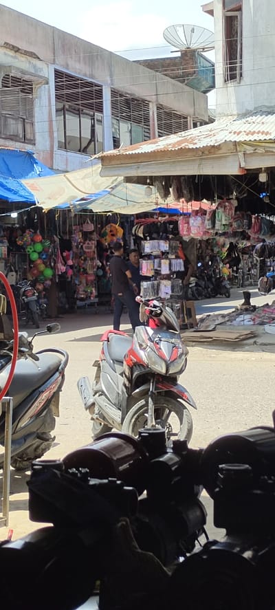 The image depicts a street scene in Lhoksukon, Indonesia. Stalls line the street, shaded by tarps and awnings. These stalls display various merchandise, including toys, bags, and small items. Two men stand near a stall, engaged in conversation. Several parked motorcycles are visible, both near the stalls and in the foreground. The backdrop features older buildings, one with a satellite dish on its roof. The foreground is dominated by blurred objects, obscuring the lower portion of the view.