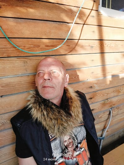 A medium shot captures a fair-skinned man with a shaved head and a light stubble beard, lying down and looking upward, with his right eye closed. He is wearing a dark jacket with a thick, light brown fur collar and a t-shirt with a graphic of a blonde woman. The man is positioned against a wooden wall, which appears to be the exterior of a structure. A green hose is draped across the upper portion of the wooden wall, casting a shadow. The overall environment suggests an outdoor setting, possibly a backyard or a rustic outdoor area. The timestamp overlaid on the image reads "realme 12 24 июня 2026 г. в 19:27". The date "24 июня 2026" translates to June 24, 2026, and the time is 19:27, indicating it was taken in the evening. The location is specified as Oseyevo, Russia.