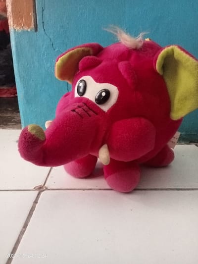 A bright pink plush elephant toy is positioned on a white tiled floor with dark gray grout lines, against a light blue painted wall. The toy features large, round ears with lime green inner linings, prominent oval eyes with black pupils on white backgrounds, a curved trunk with a lime green tip, and two small, cream-colored tusks protruding from its snout. A tuft of light-colored, matted fur is visible on the top of the toy's head. A white product tag is partially visible on the right side of the toy. The light blue wall in the background exhibits a vertical crack towards the left side, and a darker, reddish-brown object is partially visible behind the wall in the far left background. The image was captured on 2023/09/24 at 12:24 in Quebrada del Medio, Venezuela.