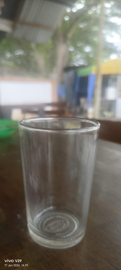 A clear glass sits on a wooden table. The glass is empty and reflects light from above. The background is blurred, showing a corrugated metal roof, trees, and some brightly colored structures. The bottom of the glass has a circular pattern embossed on it. A watermark at the bottom left reads "vivo V29 17 Jun 2026, 14.19". The scene is likely outdoors, possibly in a cafe or open-air market in Keudematangkuli, Indonesia, given the architectural elements and the watermark.