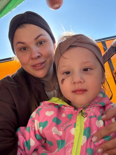 A selfie shows a woman and a toddler, likely outdoors in Кипчак, Russia. The woman wears a dark headband and a brown top. The toddler, resting against the woman, wears a pink jacket with green heart patterns and a light-brown headband. A hand with manicured nails is visible near the toddler's jacket. An orange chair and a blue sky are in the background.
