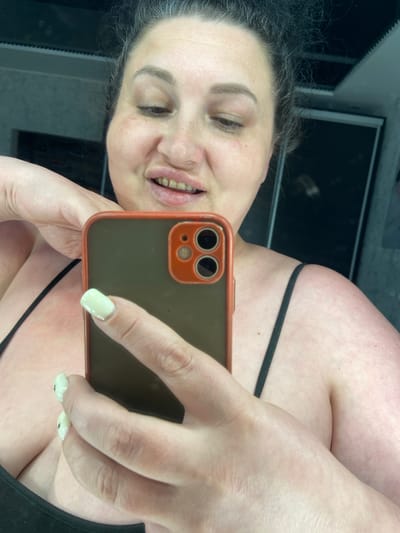 The image is a close-up, selfie-style photograph taken by a woman. She is looking into a smartphone held in her hands. Her hair is dark and pulled back. She is wearing a black camisole with thin straps. Her nails are painted a light green color. The background appears to be a bathroom with tiled walls and what looks like a shower door or partition. There is no indication of the city of Rozkvit, Ukraine, in the image.