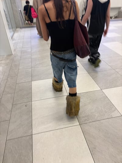 The image, taken from a high-angle perspective looking down, captures several individuals walking through a modern indoor public space, likely a shopping mall or large commercial building in Longueuil, Canada. The scene exhibits significant motion blur, particularly on the subjects, indicating movement and a dynamic shot.

In the immediate foreground, a person is visible from the chest down, wearing a black tank top or camisole, light blue cuffed jeans, and distinctive light brown/tan furry slip-on shoes or boots. A dark red or maroon shoulder bag with a fringed strap is carried across their body, and a studded or decorative belt is worn with the jeans. Long, dark, wavy hair cascades down their back. This individual appears to be walking away from the camera.

Another person is partially visible in the upper left section of the frame, largely blurred due to motion. They wear a black top and dark trousers, with dark shoes featuring light green soles. Further into the background, additional blurred figures suggest a busy environment, with one person carrying a bright pink bag and another a camouflage-patterned bag.

The floor is composed of large, alternating squares of light grey and off-white or cream-colored tiles, forming a clean and contemporary pattern. The interior appears brightly lit by artificial lighting, typical of a commercial establishment. No specific time of day is indicated, and there is no discernible text within the image.