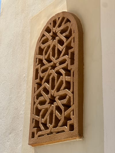 The image shows an arched, intricately carved stone window lattice set against a textured, light-colored wall. The lattice is made of reddish-brown material and features a complex geometric pattern. The wall appears to be a building facade, possibly in a warm, sunny climate given the lighting. The specific location of Ait Ouallal, Morocco, suggests a traditional architectural style often found in Moroccan towns. There are no people or animals visible in the image, nor any discernible actions or interactions. The focus is entirely on the architectural detail of the window and its surrounding wall.