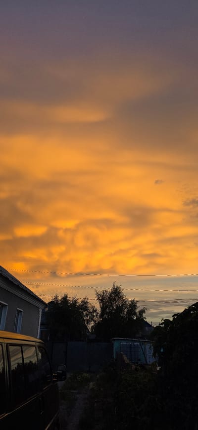 The image depicts a horizontal view of a dramatic sunset over a suburban setting in Saran, Kazakhstan. The sky is filled with vibrant, fiery orange clouds, with a gradient transitioning to a dusky purple on the left. A calm body of water is visible in the distance on the right, reflecting the colorful sky.
In the foreground, there are elements of a residential area. A portion of a building with a grey roof and white-framed windows is visible on the lower right. Adjacent to it, a dark-colored vehicle, possibly a van, is parked. Trees and dense foliage frame the scene on the right side, contributing to a sense of enclosure. Faint utility lines are also visible crossing the sky horizontally.
The time of day is clearly sunset, indicated by the intense colors of the sky. The weather appears clear apart from the dramatic cloud formations. The overall mood evoked by the image is one of awe and natural beauty, juxtaposed with the mundane aspects of a residential environment. No text is visible in the image.
