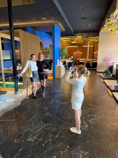 The scene takes place inside a building with a polished, dark, marbled floor. Three women are present. One woman is standing and taking a photograph with a mobile phone. Two women are posing for the photo. There is a dining or seating area visible with modern design elements in blue and gold. The location is not identifiable as Torreforta, Spain from the visual elements alone.