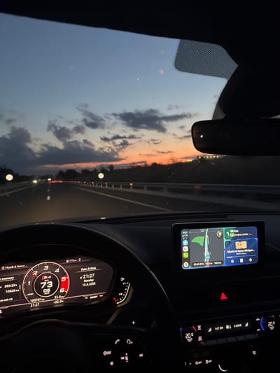 The image shows the interior of a car driving on a highway at dusk. The dashboard displays speed, engine status, and navigation information. The exterior view through the windshield reveals a highway with traffic lights and a sunset in the background. The navigation system indicates a destination of Sofia, Bulgaria, and a distance of 56 km. The time is 21:27 and the date is June 15, 2026. The car appears to be in motion, with the steering wheel visible. The overall scene suggests a journey in Bulgaria, likely approaching Ignatievo based on the navigation data.