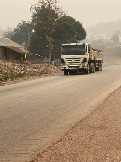 This image captures a dusty, paved two-lane road in Okura, Nigeria, under hazy atmospheric conditions. A white heavy-duty truck, bearing the registration 'KT-1AA-007' on its cab, is the primary subject, moving towards the viewer on the right side of the road. The truck features multiple rear axles and an open, empty cargo bed with high sides. To the left of the road, an unpaved, dusty shoulder slopes upward to a bank covered in accumulated waste, including plastic bags and assorted debris. Behind this bank, two rudimentary structures with rusty corrugated metal roofs are visible. The first structure, closer to the foreground, is light brown, with a person wearing a light-colored garment (possibly yellow-green) standing partially obscured nearby. Further back, another smaller, dilapidated wooden structure with a corrugated roof is present. Dense tropical foliage, including palm trees and other broadleaf trees, forms the immediate backdrop behind these structures. In the distant background, obscured by heavy haze, hills rise, dotted with scattered buildings indicating urban or semi-urban development.