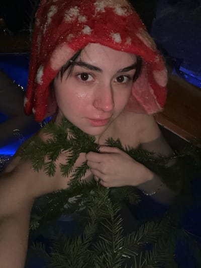 The image depicts a young adult female, partially submerged in dark blue water, likely an outdoor bath or hot tub. She has dark hair, brown eyes, and a flushed, wet face. She wears a red and white knitted or felted hat, patterned to resemble a mushroom cap, with small white particles (possibly snow or ice) visible on the hat and her hair. Coniferous branches, appearing to be fir or spruce, are draped across her chest and arms; she holds some of these branches with her right hand. A thin, light-colored bracelet is visible on her left wrist. The background is dark, suggesting a nighttime or low-light setting. The scene is located in Базарово, Russia.
