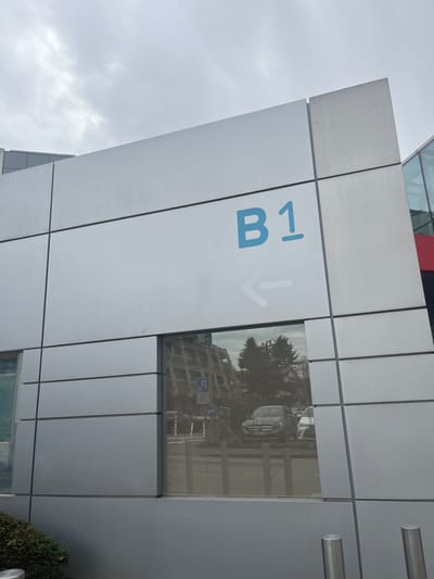 The image, rotated 90 degrees counter-clockwise from its intended viewing, depicts the exterior of a modern building in Meyrin, Switzerland, under an overcast sky. The building's facade is constructed from large, light grey metallic panels, creating a geometric, grid-like pattern.

Prominently featured on one of these panels is a sign with the text "B1" in blue, accompanied by a faint white arrow pointing downwards. This signage likely indicates a basement level or a directional cue within the building. Adjacent to the sign, a square window section reflects the surrounding environment. The reflection clearly shows the silhouette of a large tree with dense foliage, and beneath it, a multi-story building with numerous windows. Closer to the bottom of the reflection, elements resembling a yellow ladder or scaffolding and a white, possibly tent-like, structure are discernible, suggesting ongoing work or maintenance nearby. The window glass itself appears to have a fine vertical striped pattern, potentially from internal blinds or a textured surface.

The sky in the upper left corner of the image is uniformly cloudy, indicating an overcast daytime. A small, dark green bush or hedge is partially visible at the bottom right of the frame. No people are directly visible in the immediate foreground.