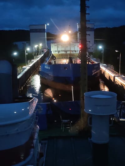 The image depicts a canal lock at night in Devyatiny, Russia. A large blue and white cargo ship, identified by Cyrillic lettering on its side as "ОКСКИЙ-69", is positioned within the concrete lock. The ship's lights are on, illuminating the water and the lock walls. The sky is dark blue with clouds, and several streetlights are visible along the lock structure, casting pools of light. In the foreground, the edge of another vessel or structure, predominantly white, is visible, along with a white cylindrical object with handles. The surrounding environment appears to be a wooded area. The overall impression is one of industrial maritime activity taking place after dark.