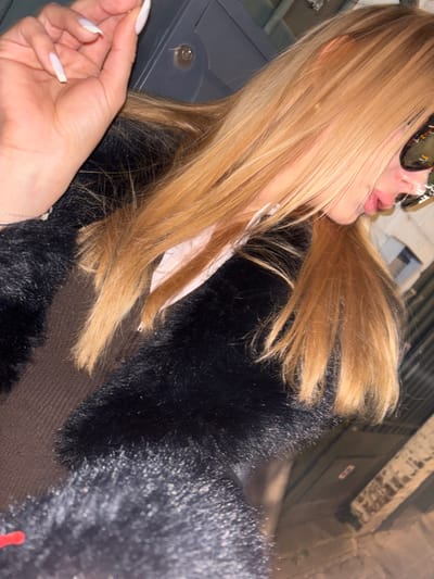 A partial portrait of a female individual is depicted in an urban setting within Asnières-sur-Seine, France. The subject has long, straight, light blonde hair. Her face is partially obscured by reflective aviator-style sunglasses, with her lips slightly parted. She is dressed in a black faux fur coat over a brown ribbed garment, with a white shirt collar visible. Her left hand is raised, revealing manicured nails with white tips, and she delicately holds a small, light-colored object, possibly a pill or fragment, between her thumb and index finger. A thin silver bracelet is on her left wrist. The background features a dark gray metallic surface with a circular keyhole or dial mechanism to the left of her head, and an indistinct, unevenly lit light-colored wall or structure to the right, suggesting an outdoor or semi-outdoor, low-light environment.